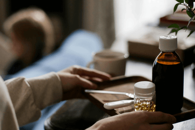 Hands hold a tray with medicine bottles, a spoon, and a thermometer. A mug and blurred background are visible.