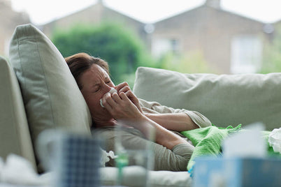 A woman sitting on a couch, using a tissue to blow her nose, displaying a moment of discomfort or illness.